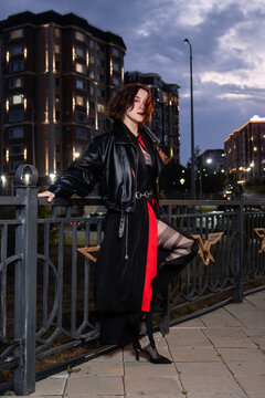 Stylish young woman in black leather jacket and black dress with red accent posing at night in urban cityscape.