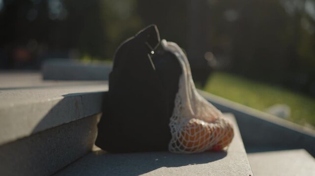 Close up of black bag with mesh net containing oranges resting on concrete steps. scene features soft natural light outdoor greenery in background and minimal camera movement emphasizing