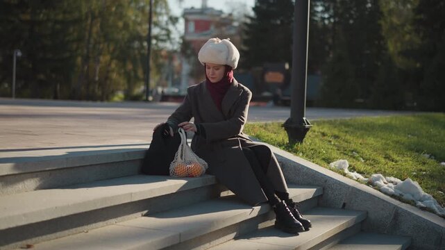 Close up of woman sitting on stone steps outdoors handling items from her bag. Woman wears winter clothing including fur hat and coat with bags containing produce nearby. scene features soft