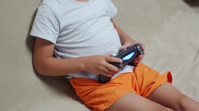 An excited child passionately holds a glowing black gamepad while playing video games. They sit cross-legged and focused on a comfortable, light-colored couch during a fun leisure routine at home.