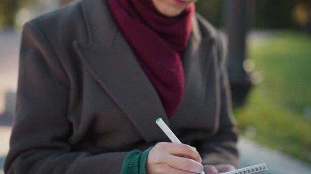Close up of woman holding pen and writing in notebook while seated outdoors. Notebook hand position coat texture visible. Soft natural light with green blurred background indicating park