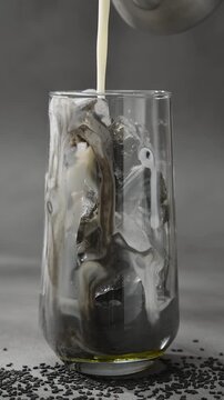 Milk pours into glass with black sesame paste and ice, creating marbled layers during iced latte preparation close up shot.