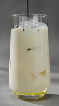 Black sesame paste pours into iced oat milk in clear glass, forming dark layer during creamy latte preparation close up.