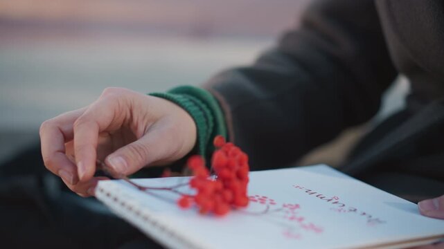 Close up of person hand holding red berries on thin stems over open notebook with handwritten text. scene features soft natural light and blurred background focusing on interaction
