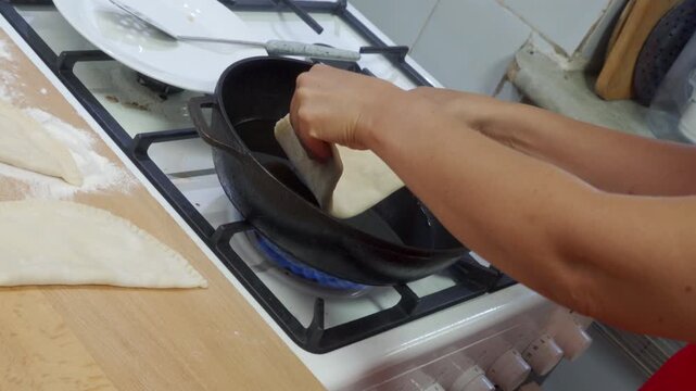 A focused cook carefully places a raw pastry into hot oil inside a frying pan. They work passionately over a lit gas stove in a bright kitchen during a joyful meal routine.