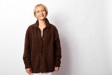 Elderly woman smiling in a studio portrait wearing a brown coat, standing on a white background,...