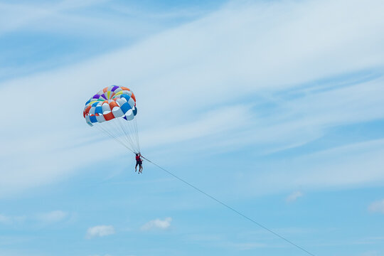 Person parasailing with colorful parachute against a bright blue sky and fluffy white clouds, showcasing an adventurous outdoor activity. water sports concept