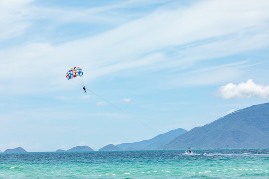 Person parasailing above turquoise ocean waters with colorful parachute, mountains visible in the background under a clear blue sky. water sports concept