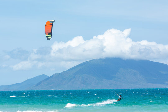 Kite surfer riding waves on a sunny beach with colorful kite soaring above, mountains and clouds in the background. water sports concept