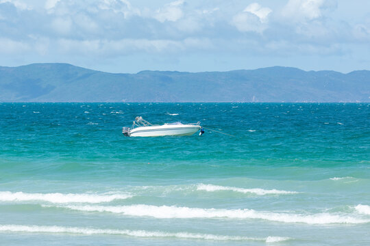 Small motorboat floats in clear turquoise waters, anchored with a buoy, while gentle waves lap at the shore. Distant mountains rise under a partly cloudy sky, creating a serene coastal scene