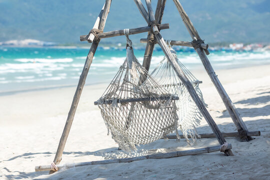 Hammock made of netting is suspended between wooden posts on a sandy beach, with turquoise ocean waves gently lapping at the shore and distant mountains. Summer tropical vacation and relax concept