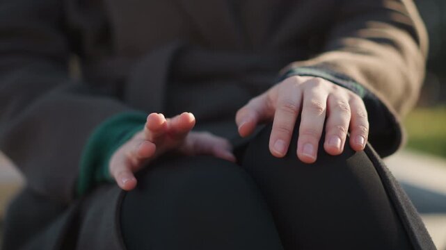 Close up of woman hands resting on her knees with fingers slightly spread showing natural skin texture and relaxed posture. subject wears dark pants under brown coat sleeve suggesting casual