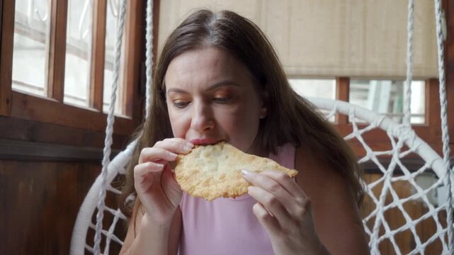 A pleased woman eagerly bites into a large, crispy golden pastry. She sits contentedly in a white hanging basket chair on a rustic wooden terrace during a cozy snack break.