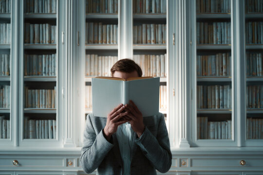 Male reader holding a book in front of his face, standing in a library with shelves filled with books, creating an atmosphere of knowledge and discovery