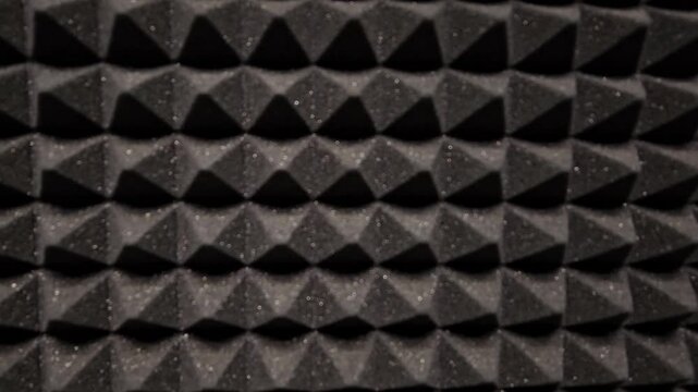 Close-up of black triangular acoustic foam texture. Dark geometric pattern fills entire frame. Surface shows soft, porous, repeating triangular shapes