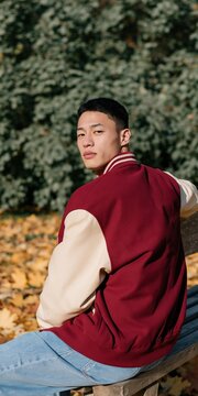 Young man burgundy varsity jacket sitting on park bench among autumn leaves for lifestyle portrait background and casual fashion imagery