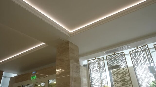 Low-angle interior view of ceiling corner with recessed cove lighting, crown molding, polished stone column, and backlit patterned screens in upscale lobby, with ceiling, lighting, molding, crown