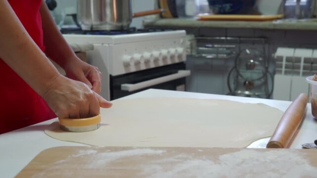 Close view of hands pressing round cutter into rolled dough on floured countertop in home kitchen, with warm overhead light, pastry texture, scattered flour, rolling pin, stove, and baking setup