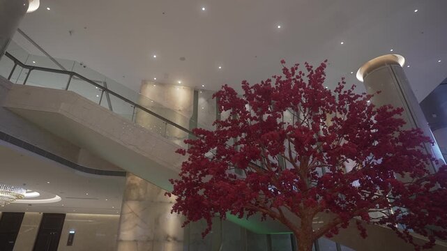 Low-angle view of upscale indoor atrium with marble staircase, glass balustrade, recessed ceiling lights, and tall red blossom tree creating elegant modern architecture with spacious lobby atmosphere