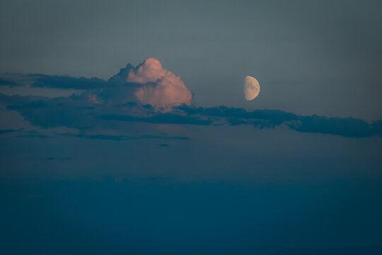 Moon partially illuminated in a clear sky with soft clouds illuminated by sunset hues, creating a serene twilight atmosphere over the landscape