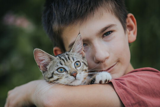 Young boy with short dark hair holds a gray tabby cat close to his face, outdoors with blurred greenery in the background, showcasing a bond between child and pet