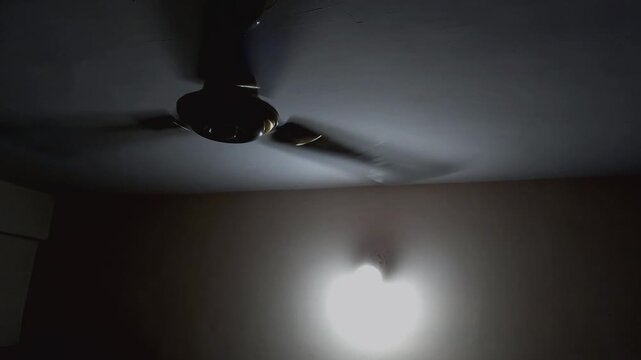 A minimalist fan rotates in a darkened room with closed blinds, showcasing passive cooling techniques used to survive extreme heat without overloading the electrical grid.