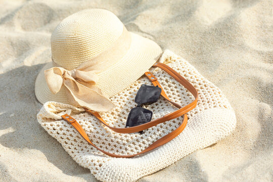 Straw sun hat with ribbon, black sunglasses, and woven beach bag resting on sandy surface under bright sunlight. Summer outfit and vacation concept. Copy space. Top view