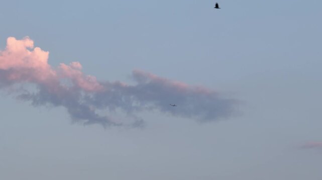 A small airplane silhouette crosses a hazy dusk sky, flying toward a visible moon partially obscured by drifting clouds. The minimalist composition captures the quiet vastness of air travel