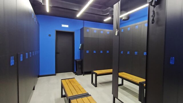 Blue walls surround dark lockers with numbered doors. Wooden benches sit beside lockers, ready for users. Bright ceiling lights illuminate the clean, modern space