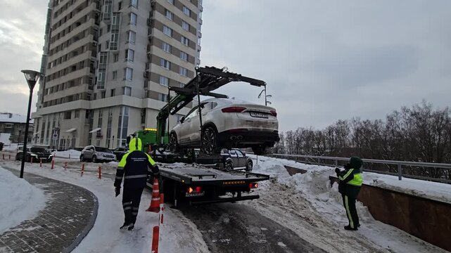 MOSCOW - FEB 8, 2026: Snowy street with tow truck lifting a white car. Workers in bright vests manage the scene. Modern building stands tall behind the scene. Traffic cones mark the area around the