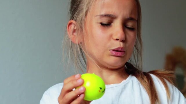 Young girl with long hair holds squishy green dumpling toy close to her nose, reacting to its scent in a bright indoor setting with neutral background