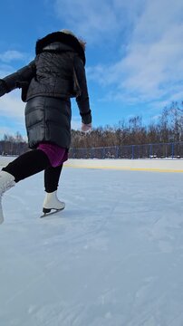 Lone skater glides across an outdoor ice rink. Cold winter sky frames her silhouette against sunlit clouds. She wears a dark coat and skates with quiet confidence