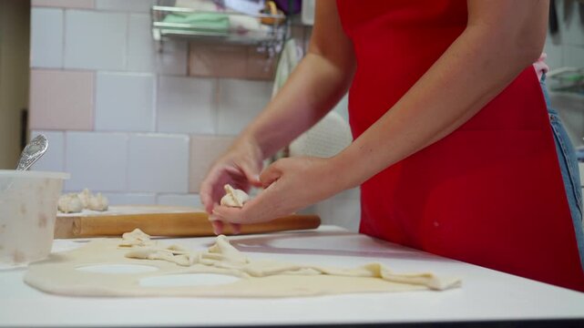 Close-up of woman shaping homemade dumplings on floured kitchen counter, hands pinching dough wrappers with filling, red apron visible, tiled backsplash and warm light adding domestic cooking