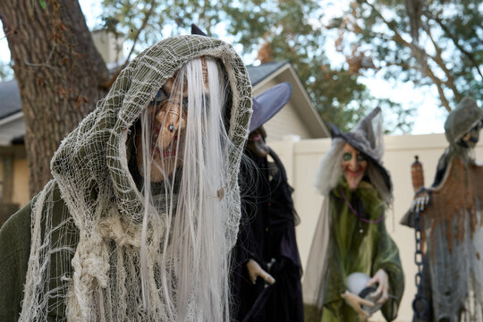 Creepy hooded figure with white hair and tattered gauze leans toward the camera. Halloween yard decor with other witches blurred in the background.