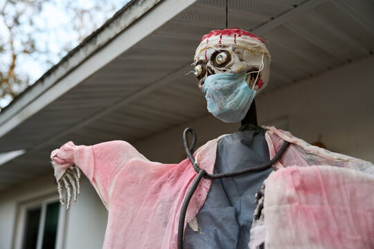Low-angle view of a Halloween zombie doctor prop with bulging eyes, blue mask, and bloody bandage. Creepy medical-themed yard decoration on a house porch in daylight.