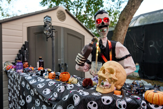 Red-eyed skeleton bartender pours a drink behind a skull-patterned table. Spooky outdoor Halloween party setup with props, pumpkins, and candy.