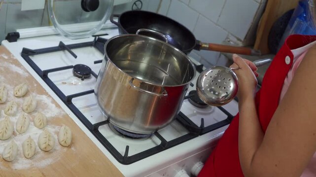 Close view of person wearing red apron ladling hot water from stainless steel pot on gas stove beside tray of raw dumplings; bright kitchen lighting, white tiled backsplash, metal ladle, stovetop