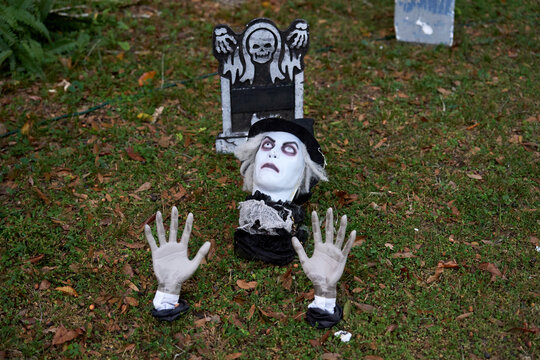 A pale ghost head and two hands emerge from the ground in a graveyard beside a tombstone. Spooky Halloween cemetery decoration with vintage costume and eerie eyes.