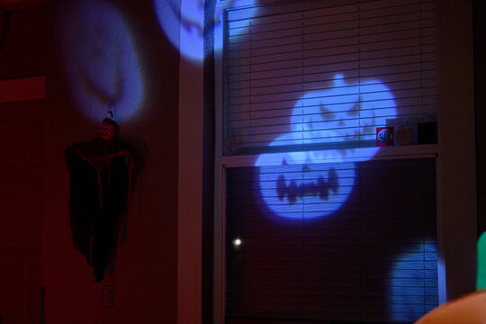 Blue pumpkin face is projected onto a house window at night. Halloween home decoration with spooky light and blinds.