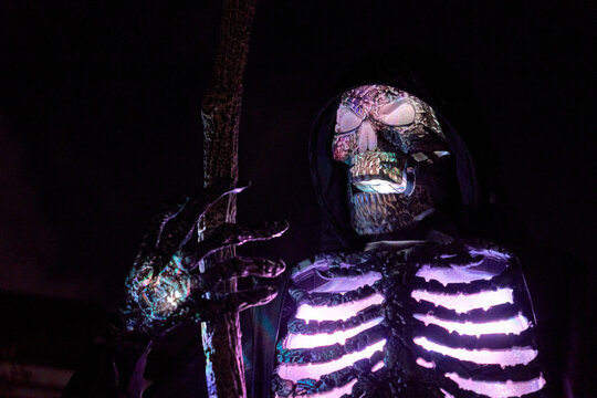 Hooded skull figure with staff shows shimmering LED ribs at night. Multicolor glow lights a spooky Halloween decoration.