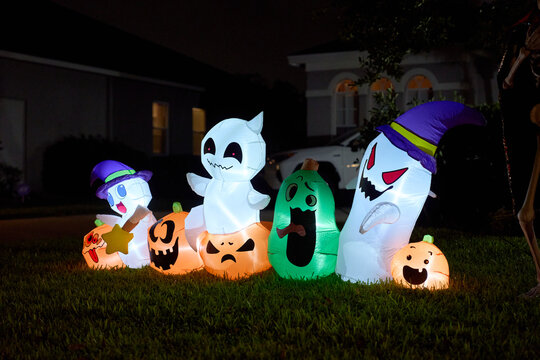A lineup of smiling ghosts and carved pumpkin inflatables lights a suburban yard. Festive Halloween scene with vivid colors after dark.