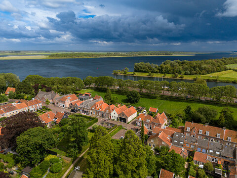 High angle wide horizontal aerial drone photograph of historic orange roofs, Lake Veere shoreline, green polder landscape, and dramatic storm sky in Veere, Zeeland, Netherlands on sunny summer day