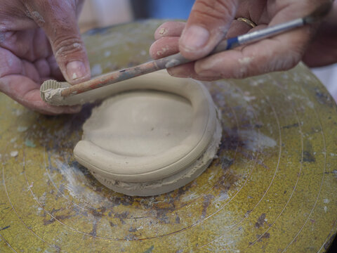 An artisan carefully pressing and shaping a circular raw clay base on a studio table focusing on traditional handbuilding techniques and the fundamental process of authentic ceramic craftwork.