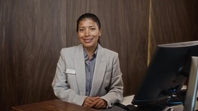 Portrait of Hispanic young adult woman working as receptionist, smiling at camera behind hotel lobby desk. Clip is perfect for hospitality marketing and customer service training videos
