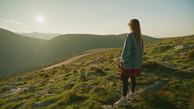 Person stands on a mountain slope looking at the sunset while wearing a denim jacket and black leggings