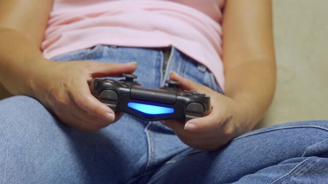 Closeup view of person seated on couch holding illuminated game controller with both hands, thumbs on analog sticks and index fingers near shoulder buttons, wearing blue denim jeans and light pink