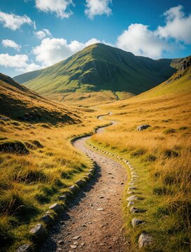 Winding dirt path goes through grassy valley toward green mountain under blue sky. Stones line path side. Rural landscape offers wide open outdoor space.