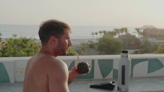 Young man drinking traditional yerba mate from a calabash gourd on a balcony overlooking the ocean