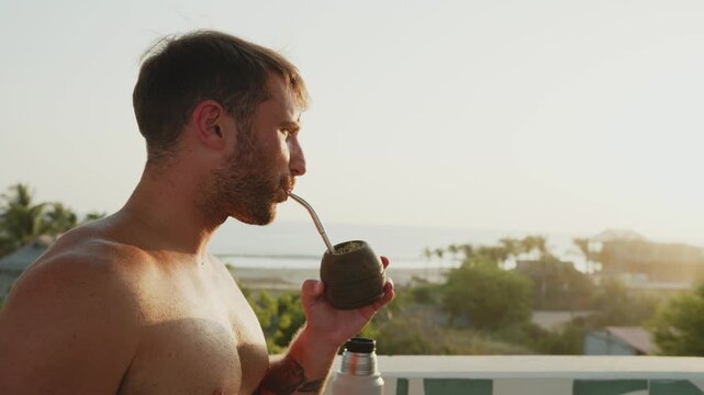 Young man enjoying a hot yerba mate tea while looking at the ocean view from a balcony at sunrise