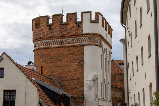 Medieval brick gate tower from 1432 stands beside historic buildings in Torun. Crenellations, patterned masonry and a steep tiled roof create a clear Gothic layout for heritage.
